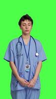 Front view Young nurse with medical expertise poses with confidence, wears blue scrubs uniform and smiling against greenscreen backdrop. Health assistant gives support to doctors as profession. Camera B. video