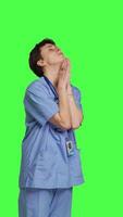 Side view Hospital nurse worshipping God by prayer against greenscreen backdrop, praying to jesus christ and having hope in christianity and spirituality. Medical assistant doing meditation. Camera B. video