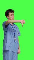 Side view Displeased medical assistant shows thumbs down in studio, presenting her disapproval and disagreement with dislike symbol against greenscreen. Nurse feeling unhappy with rejection sign. Camera B. video