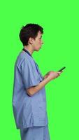 Profil jung Krankenschwester SMS auf Smartphone gegen grüner Bildschirm Hintergrund, mit Handy, Mobiltelefon Telefon zu prüfen Sozial Medien Mitteilungen auf Arbeit Unterbrechung. medizinisch Assistent Surfen online Netz Seiten. Kamera b. video