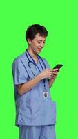Side view Young nurse texting on smartphone against greenscreen backdrop, using mobile phone to check social media messages on work break. Medical assistant browsing online web pages. Camera B. video