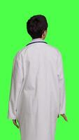 Back view Woman physician looking around for patients to come at checkup exams, waiting for someone standing against greenscreen backdrop. General practitioner with white coat feeling impatient. Camera B. video