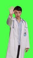 Front view Woman medic waving hello and asking people to come over at checkup, inviting patient to come closer and greets them. Skilled physician with white coat and stethoscope against greenscreen. Camera B. video