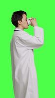 Profile Woman physician drinking a cup of coffee against greenscreen backdrop, taking a break from medical work. Specialist serving caffeine refreshment in studio, wearing a white coat. Camera B. video