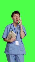 Front view Nurse answering landline phone call to make checkup appointments, talking to patients on telephone with cord against greenscreen backdrop. Health specialist in scrubs uses vintage phone. Camera B. video