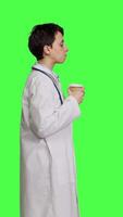 profiel gelukkig medisch portie heet koffie kop net zo verfrissing tegen groene scherm achtergrond, vervelend wit jas en drinken cafeïne drank. algemeen beoefenaar genieten van drankje, medisch expertise. camera b. video