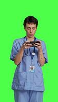 Front view Medical assistant playing mobile video games on smartphone app, taking a break from healthcare work. Nurse enjoys gaming tournament on phone, standing against greenscreen backdrop. Camera B.