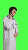 Side view Woman doctor talking on smartphone call against greenscreen backdrop, answering phone line to make an exam appointment for a patient. Physician uses mobile phone to chat with people. Camera B. video