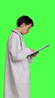 profil läkare tar anteckningar efter patient undersökning mot grönskärm bakgrund, skrivning ner läkemedel till bota sjukdom. läkare i vit täcka använder sig av Urklipp papper för kolla upp. kamera b. video