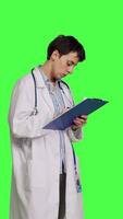 Side view Physician taking notes after patient examination against greenscreen backdrop, writing down medicaments to cure illness. Doctor in white coat using clipboard papers for checkup. Camera B. video