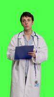 Front view Physician taking notes after patient examination against greenscreen backdrop, writing down medicaments to cure illness. Doctor in white coat using clipboard papers for checkup. Camera B. video