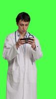 Front view General practitioner playing video games on mobile phone app, having fun with online gaming competition against greenscreen backdrop. Cheerful doctor relaxing with internet game. Camera B.