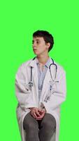 Front view General practitioner acting impatient sitting on a chair against greenscreen backdrop, waiting for patients to attend checkup appointments. Woman specialist in white coat waits for people. Camera B. video