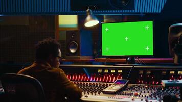 African american music producer working with greenscreen on pc, using mixing console to do mix and master in control room. Audio technician recording tracks with knobs and switchers. Camera A. video