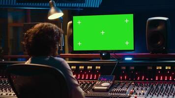 Sound designer watches online tutorials via greenscreen monitor to mix and master tracks and create music for his album. Audio engineer producing songs by twisting knobs and pushing faders. Camera A. video