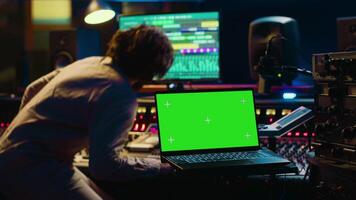 Audio technician leveling sound on recorder mixing console, having a laptop that shows greenscreen display. Producer turns up the volume of tracks in professional studio, control room setup. Camera A. video