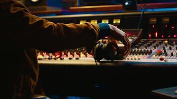 African american music producer recording tracks and editing them in post production, using mixing console with knobs and sliders. Audio expert processing sounds with technical gear. Camera A. video