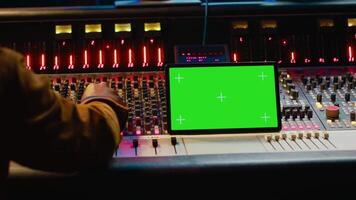 Music producer holding a tablet with greenscreen display on mixing console in control room, twisting knobs and pushing faders to edit audio recordings. Processing sounds. Camera A. video