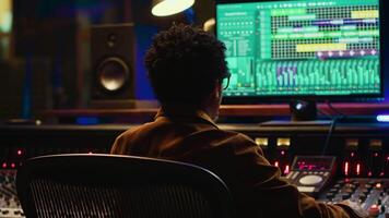 African american music producer mixing and mastering tracks on audio console, twisting knobs and pushing sliders to edit tunes. Sound engineer producing new songs in control room. Camera A. video