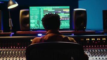 African american sound producer editing tracks on audio software and console, adjusting volume levels and adding effects in post production. Technician producing new hit song in studio. Camera A. video