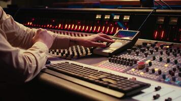 Audio technician uses mixing console and faders in control room, remote control for adjusting sound on mixer. Music producer changes volume level, creates tracks with technical equipment. Camera B. video