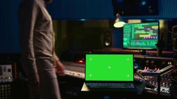 Audio technician leveling sound on recorder mixing console, having a laptop that shows greenscreen display. Producer turns up the volume of tracks in professional studio, control room. Camera B. video
