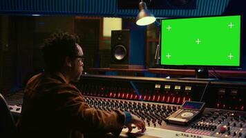 African american sound designer looks at greenscreen to record and edit tunes, uses mixing console to create new songs. Skilled technician works on processing and track recording. Camera B. video