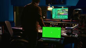 Music producer recording tracks next to laptop showing mockup isolated display, working with mixing console and mastering techniques in control room. Audio engineer producing new tunes. Camera B. video