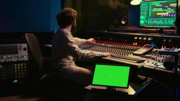 Sound designer processing and recording tracks in control room, using mixing console and sliders to adjust volume level. Technician working with greenscreen tablet in professional studio. Camera B. video