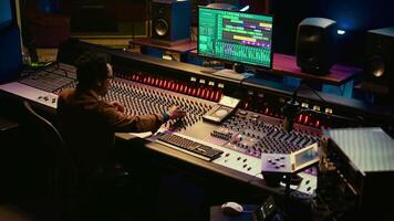 Portrait of professional sound engineer recording tracks and editing tunes by adjusting volume levels with knobs. Producer creating music with mixing console in post production studio. Camera B. video