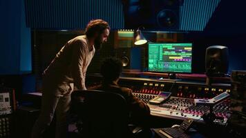 Tracking engineer collaborating with musician to edit sounds in studio control room, recording tunes to create a new album. Audio expert operating audio mixer and controller. Camera B. video