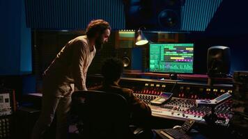 Tracking engineer collaborating with musician to edit sounds in studio control room, recording tunes to create a new album. Audio expert operating audio mixer and controller. Camera B. video