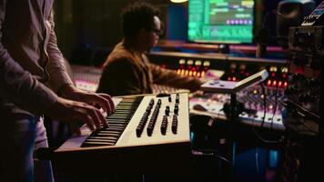 Musician playing piano midi controller in control room at professional recording studio, creating sounds with electronic keyboard notes. Sound engineer does mix and master on files. Camera B. video