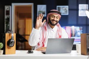 Sitting at his workplace, an Islamic guy waves at his digital laptop, starting a video conference. He is engrossed with technology, using his minicomputer to explore, interact, and conduct research. photo