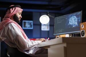 Islamic man uses AI and machine learning to ensure cyber security for a network. He monitors data centers and servers working with a PC monitor displaying advanced software and a neural network. photo