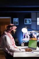 Arab male professional wearing traditional attire is seated at an office desk, using a laptop with an empty green screen. The image portrays a modern workspace for advertising or presentations. photo