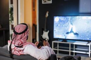 Arab man is concentrated and immersed, he controls the gameplay using a console controller. The digital world comes alive on his television display as he grasps wireless technology. photo