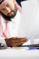 Selective focus on papers being held by a young man dressed in Arabic clothing and having a phone call. Close-up of a Muslim guy focused on examining his paperwork while speaking on a mobile device. photo