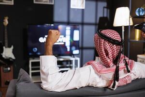 Detailed image of Arab male person celebrating his victory with fist in the air while seated on comfy couch. Rear-view shot of Middle Eastern man grasping a wireless controller. photo