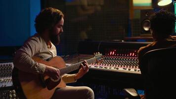 Musician and sound engineer recording guitar tunes in control room at professional music production studio. Artist playing electro acoustic instrument, live performance in soundproof space. Camera B. video