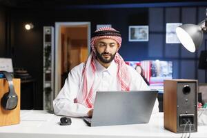 árabe chico enfocado en su ordenador portátil eficientemente surf el Internet. joven hombre vistiendo islámico ropa encarna productividad en el digital era, utilizando su personal computadora en el mesa. foto