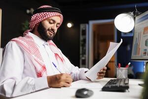 Islamic man working at his desk, surrounded by a desktop computer, documents, and paperwork, showcasing a busy corporate workspace. Side-view shot of Muslim guy writing down notes from research. photo