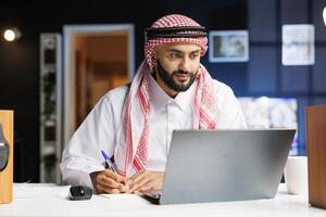 A focused Arab entrepreneur efficiently works on his laptop at a well-organized office desk. He types, browses the internet, and takes notes while surrounded by modern technology. photo