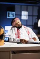 Muslim entrepreneur in traditional attire, speaking on his cellphone and checking notes on a laptop. Arab businessman conversing with a coworker and comparing research on a minicomputer. photo