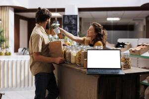 Vendor showcasing products in zero waste marketplace venue, using mockup laptop to advertise her business. Farmer trading healthy food items during local event using copy space on device for promotion photo