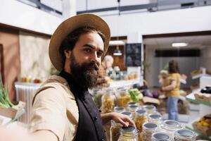 Smiling zero waste shop entrepreneur using selfie camera on cellphone to promote environmentally friendly vegan products online. Low carbon footprint supermarket owner filming marketing content photo