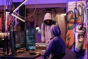 Hacker in hood showing black box with hacking technology to break into server database. Young criminal opening case with equipment for online cyberattack in abandoned warehouse photo