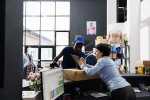 African american courier discussing shipping details with employee, checking online order in on tablet computer. Store worker standing at counter desk, preparing packages for delivery in boutique photo