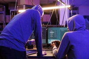 Two hackers running malicious software on computer screen to breach secure data. Criminals group wearing hoodies while breaking law and working together in abandoned warehouse photo