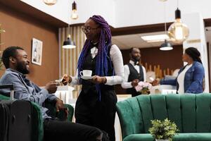 Hotel waitress holding cup of freshly brewed coffee taking cash payment from guest while serving hot drink. Happy African American man traveler ordering espresso while waiting in lobby for check-in photo